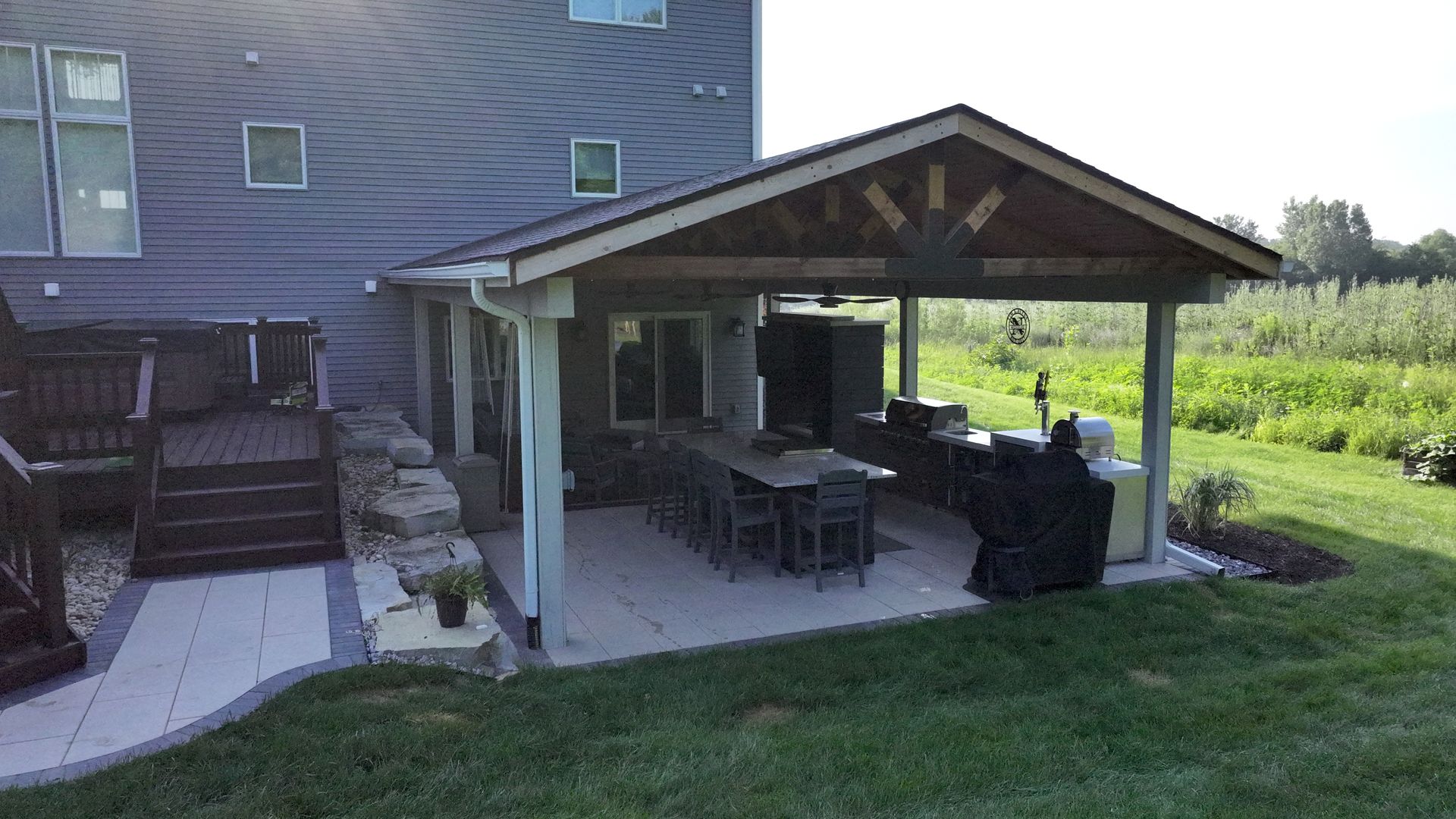 Backyard patio with dining area, kitchen, and roof. Green grass and modern home in the background.