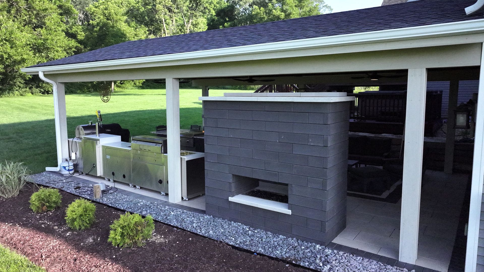 Outdoor kitchen under a roof, featuring a gray brick structure, stainless steel appliances, and a green lawn.