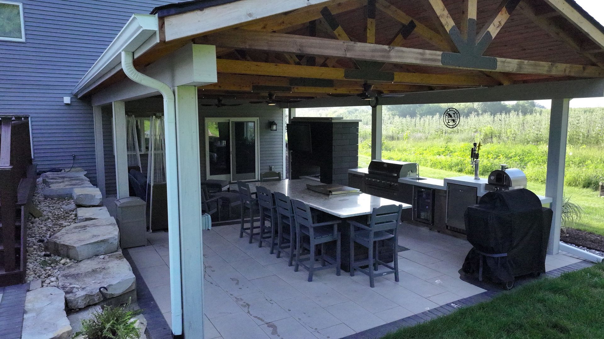 Covered outdoor kitchen and dining area with a long table, grill, and stone retaining wall.