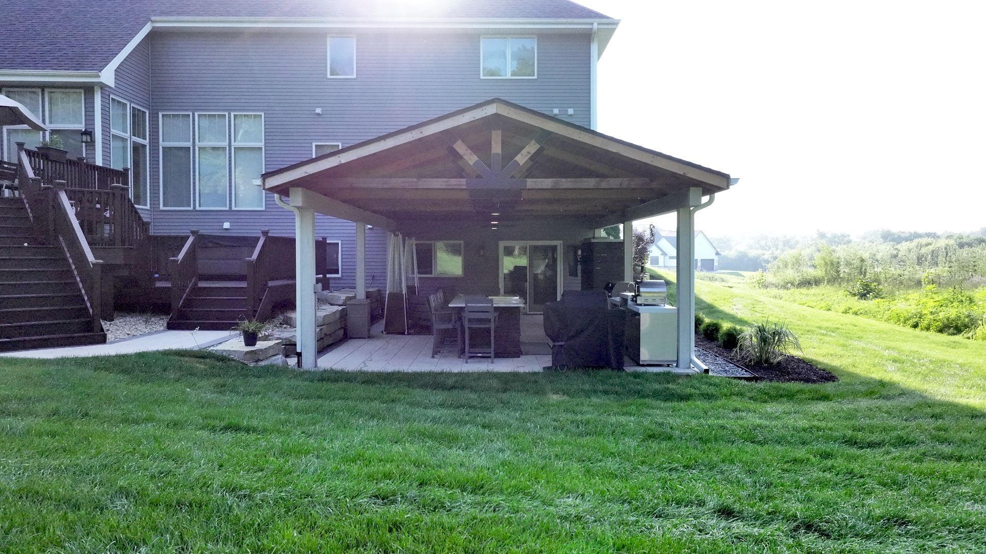 Backyard with covered patio, grill, seating, and house with deck, green lawn.
