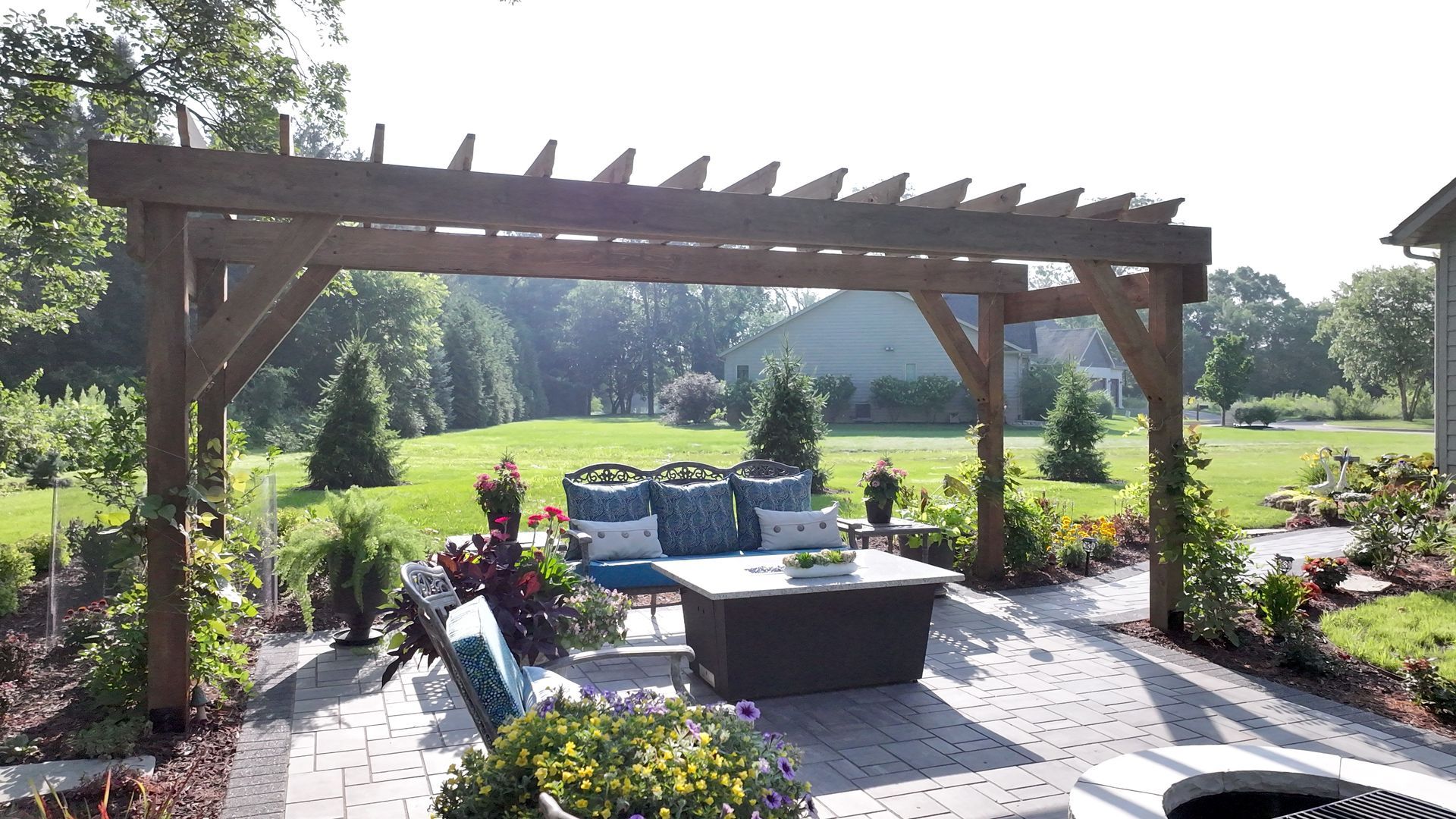 Pergola over patio with seating area; lush green lawn and trees in the background.
