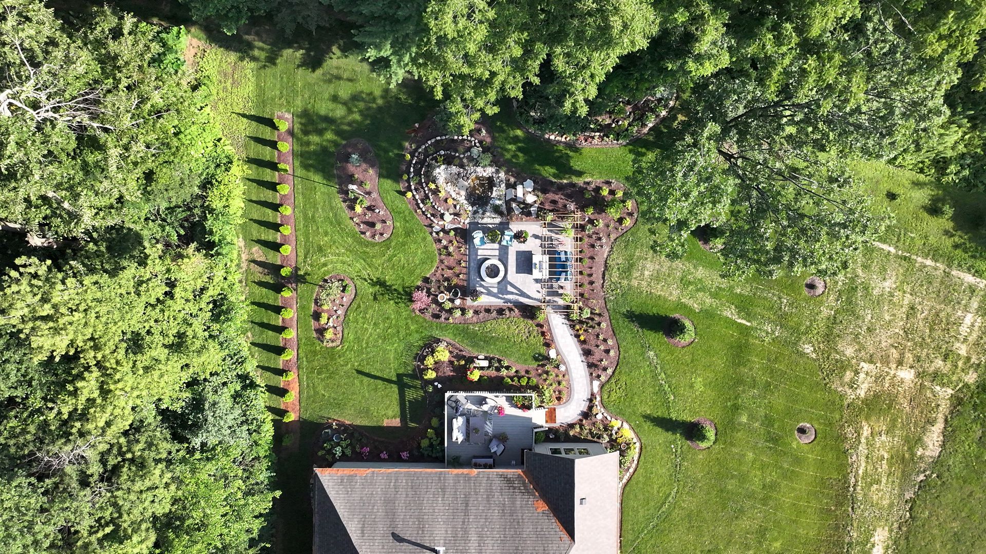 Overhead view of backyard patio with seating area, fire pit, and gardens, surrounded by trees.