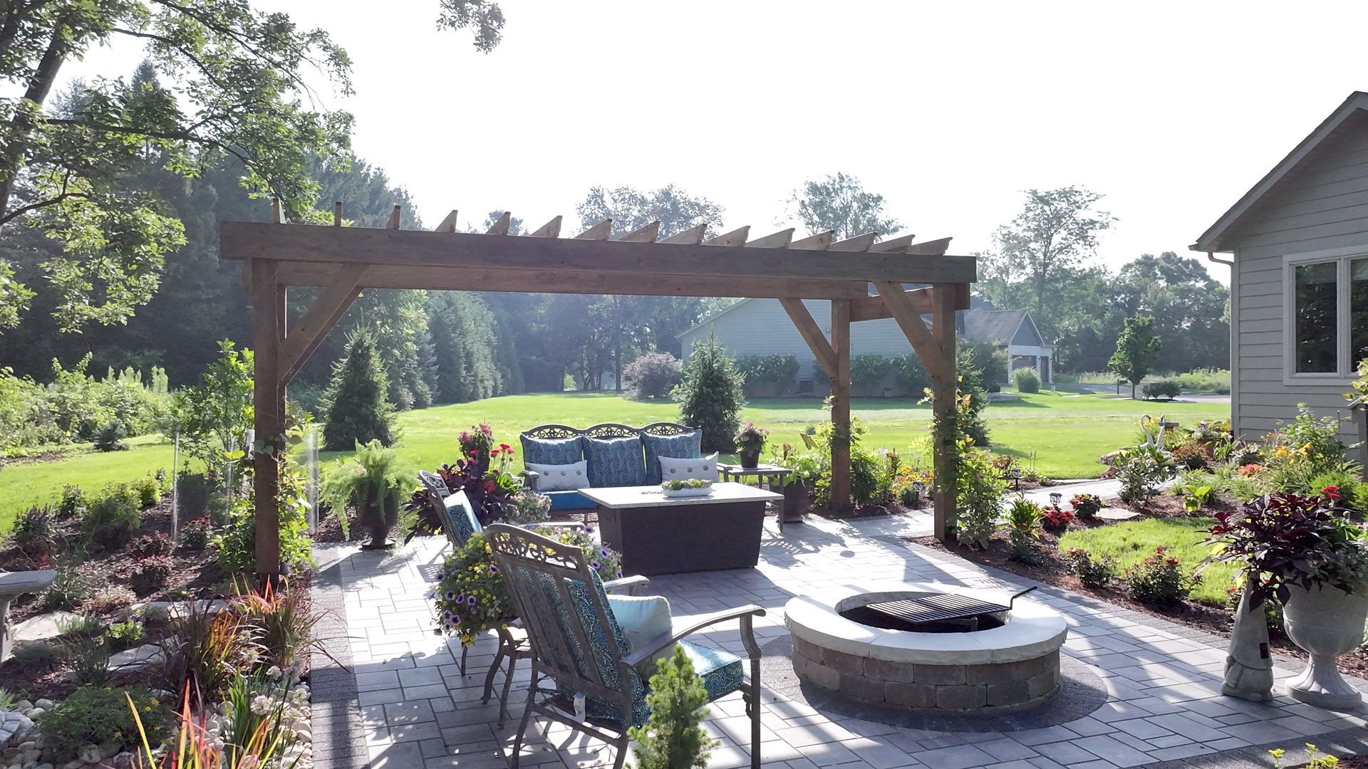 Outdoor patio with wooden pergola, seating area, fire pit, and lush green yard.
