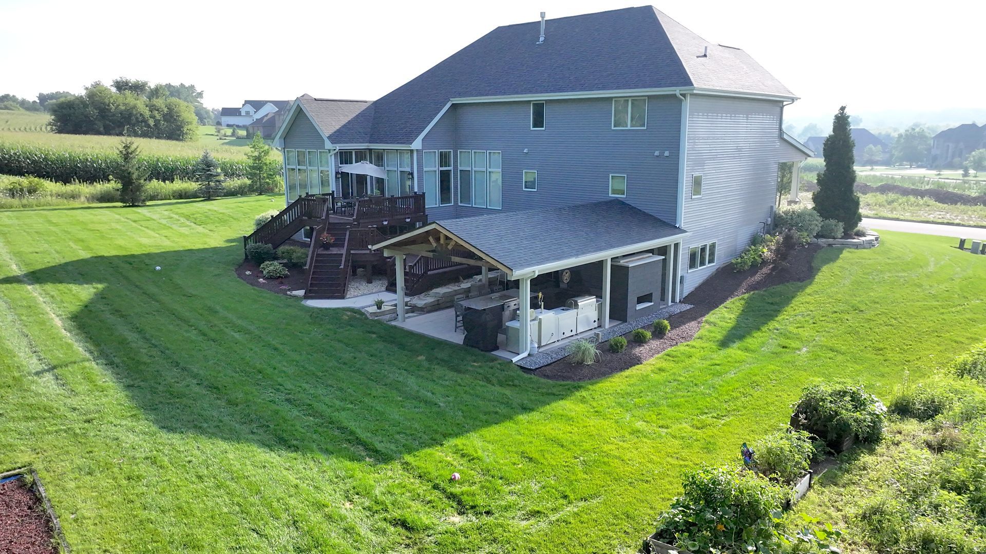 Two-story blue house with a large deck and covered patio on a grassy hill, with a view of fields.