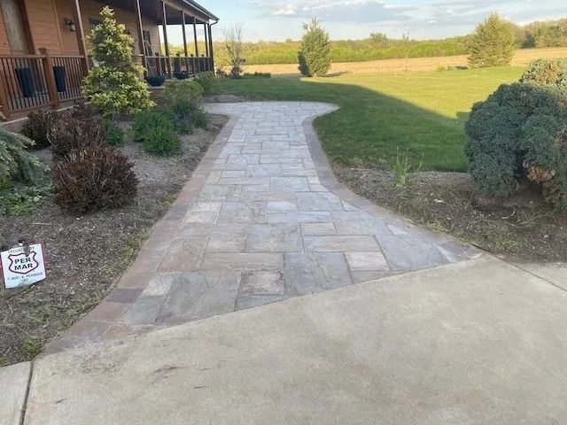 A brick walkway leads to a house with a porch, flanked by landscaping on a sunny day.
