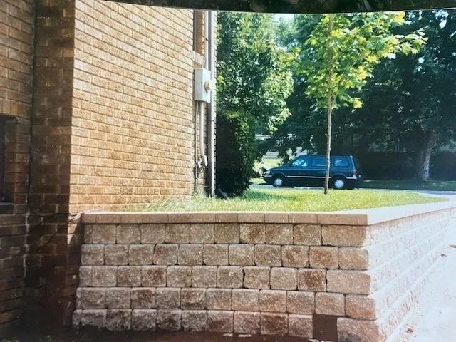 Brick retaining wall beside a building, with a car parked in the background near trees.