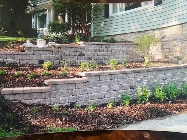 Multi-tiered retaining walls of gray blocks with plants and mulch in front of a house.