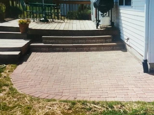Brick patio with steps leading to a wooden deck, beside a white house.