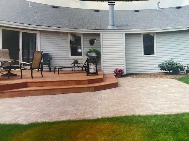 Backyard with deck, patio, house. Wooden deck with chairs, grill, curved house with windows, and a brick patio.