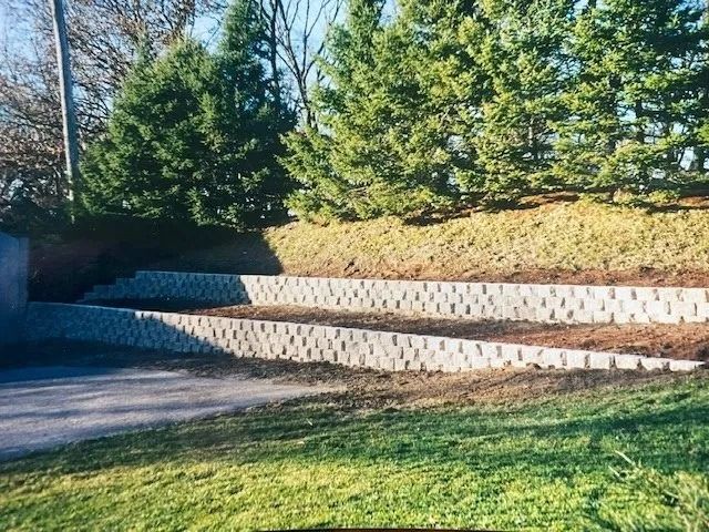 Two-tiered retaining walls made of blocks, holding back a hillside with evergreen trees.