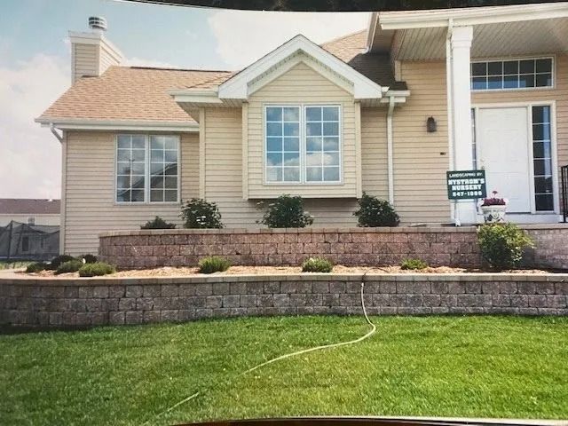 Beige house with a bay window and white columns, in front of a brick retaining wall and green lawn.