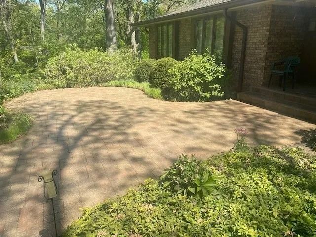 Brick patio surrounded by green bushes and trees, next to a brick house.