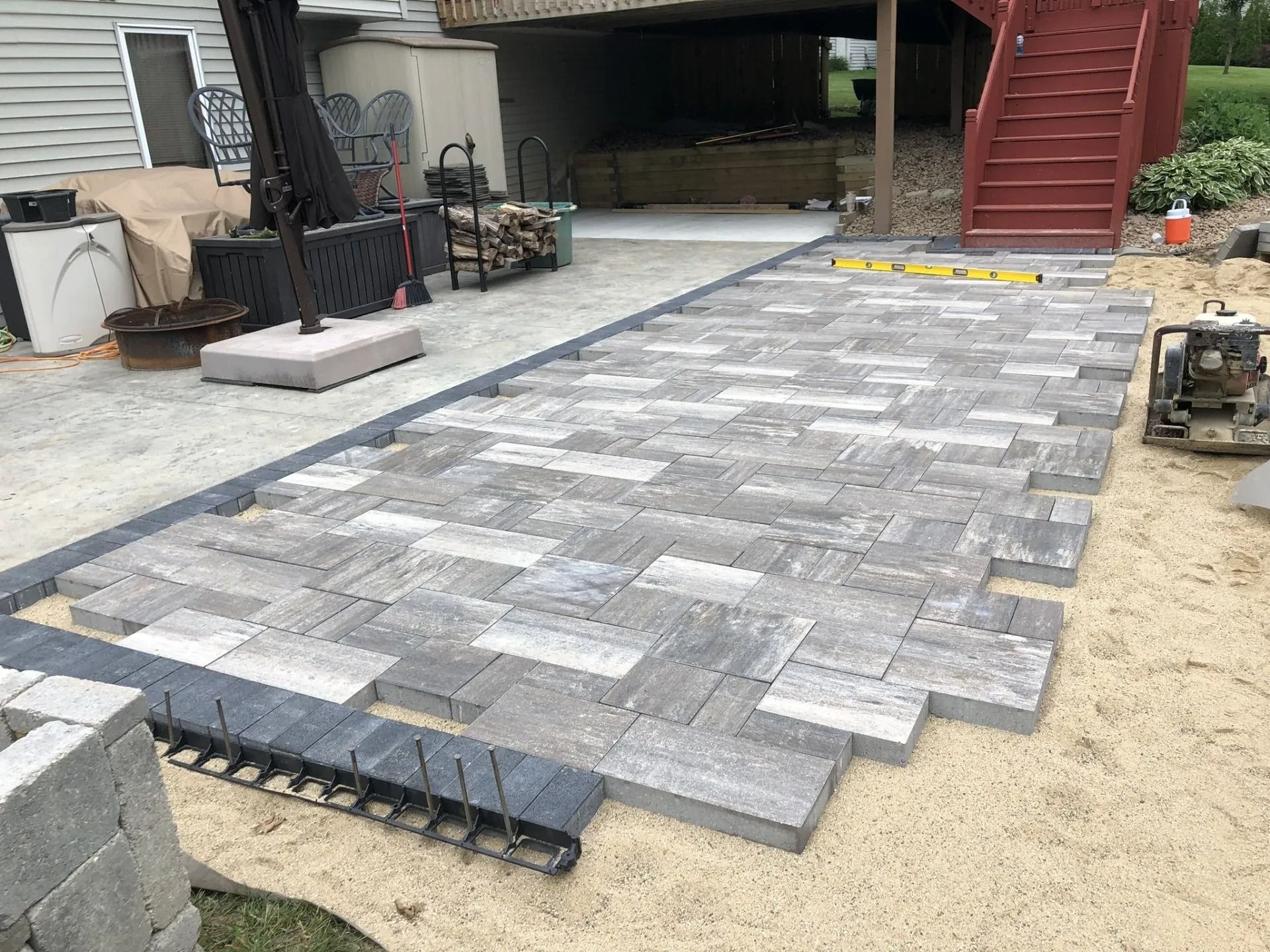 Pavers being laid in a herringbone pattern on a sand base.  Outdoor setting with stairs and a deck.
