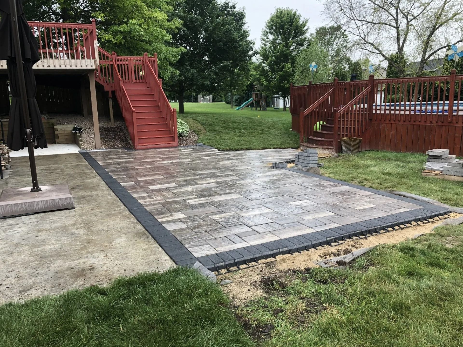 Newly constructed brick patio with dark border, adjacent to a wooden deck and backyard with grass and trees.