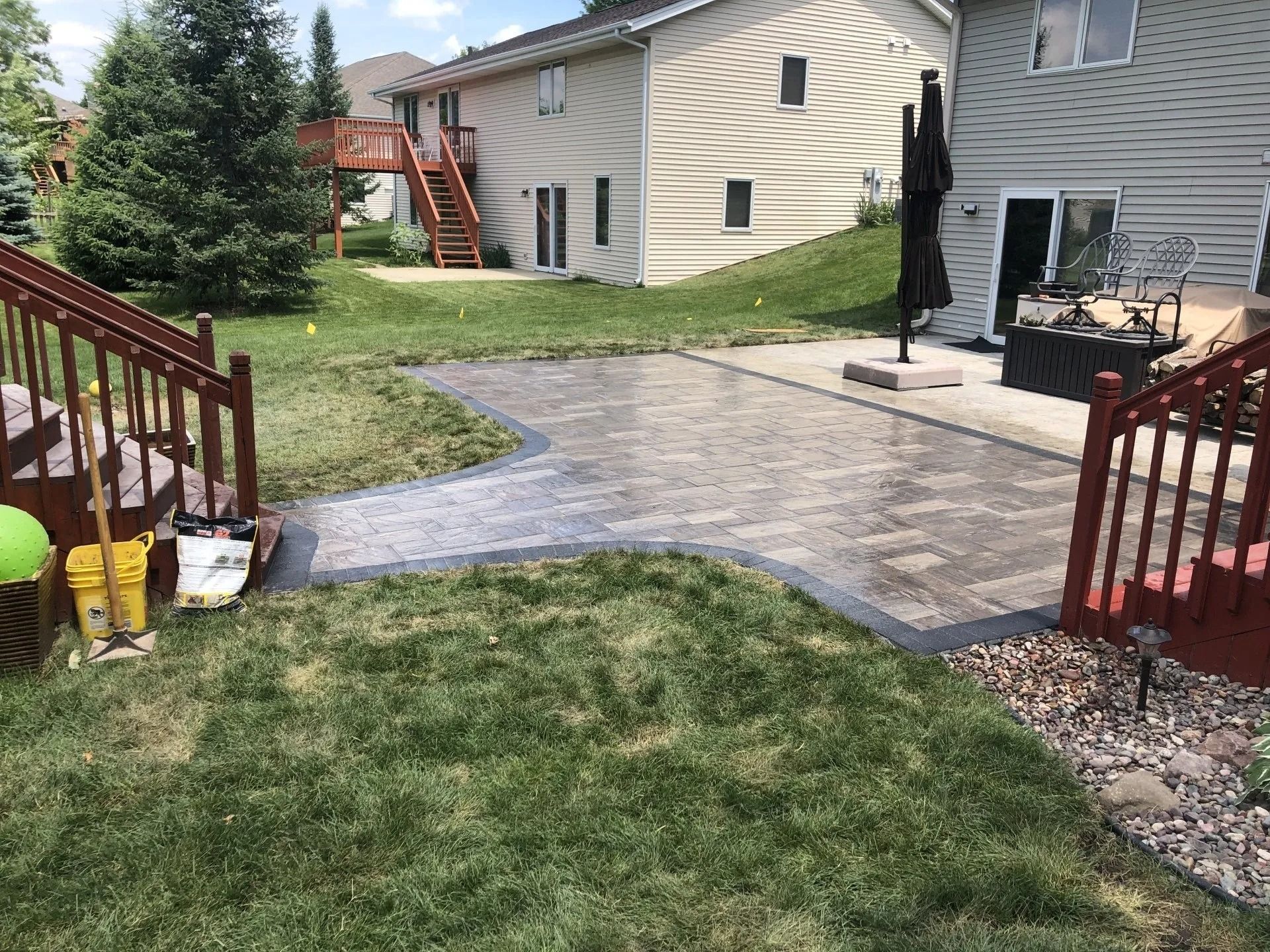 A brick patio with dark border in a backyard, between grass and deck of a house.