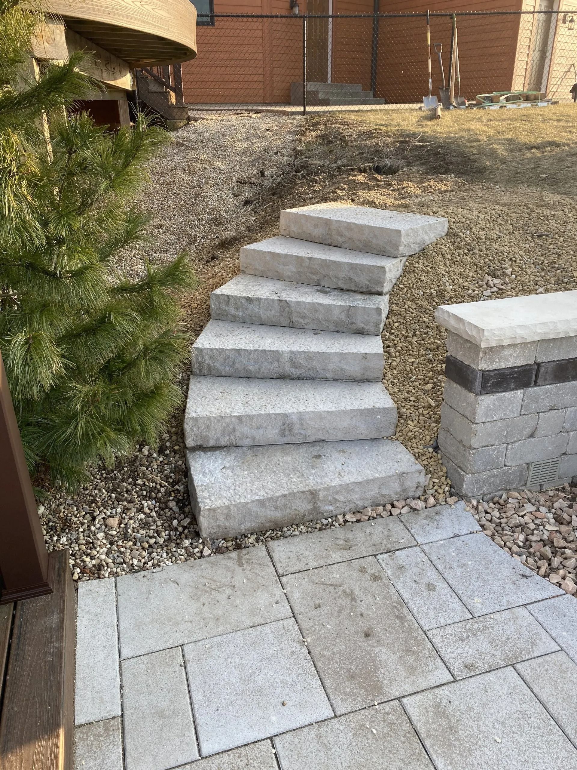 Stone steps curve up a slope in a yard, leading from a patio to a higher level with mulch and sparse vegetation.