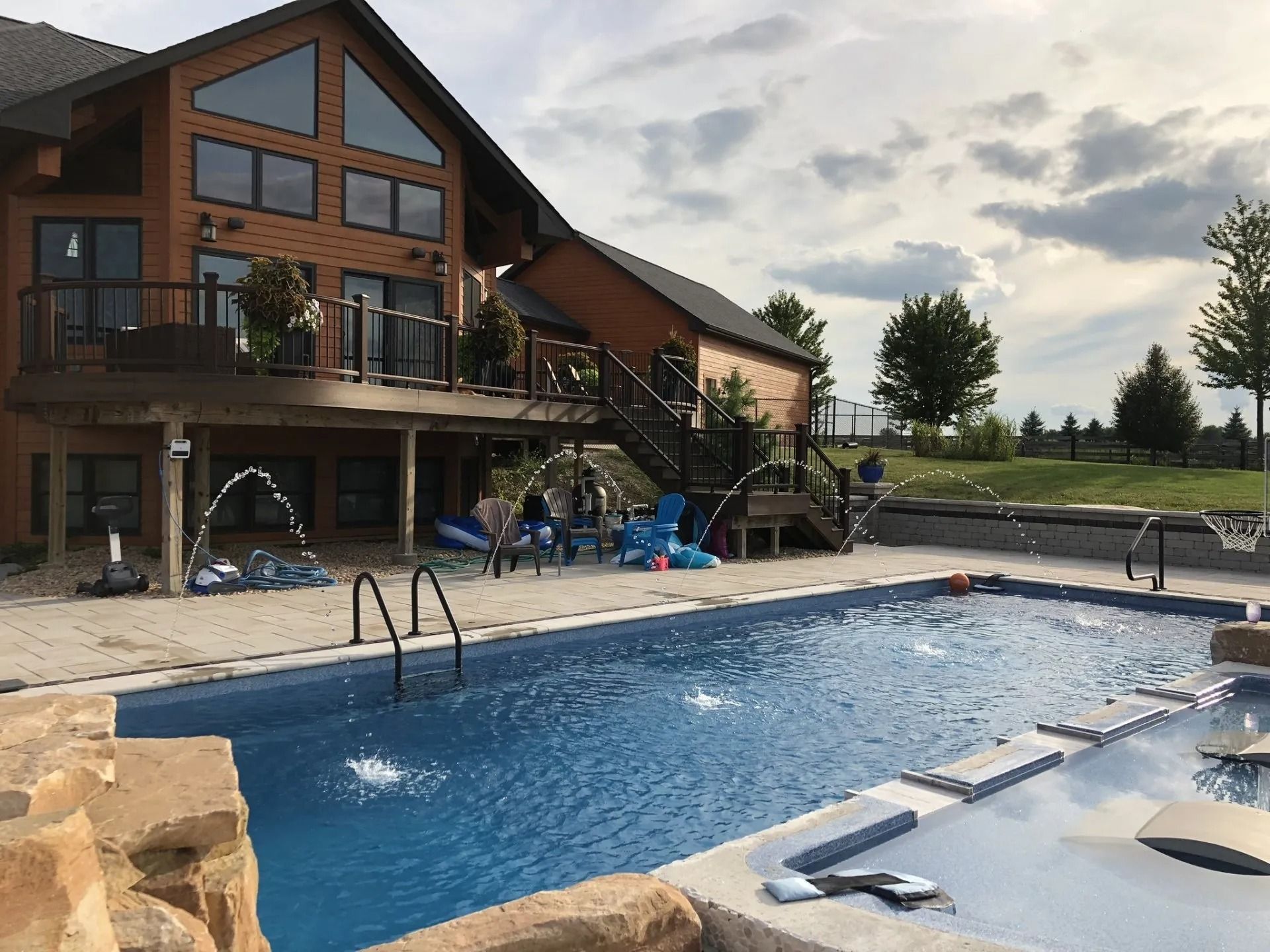 A house with a pool. The blue pool has fountains. Patio chairs and landscaping are visible.