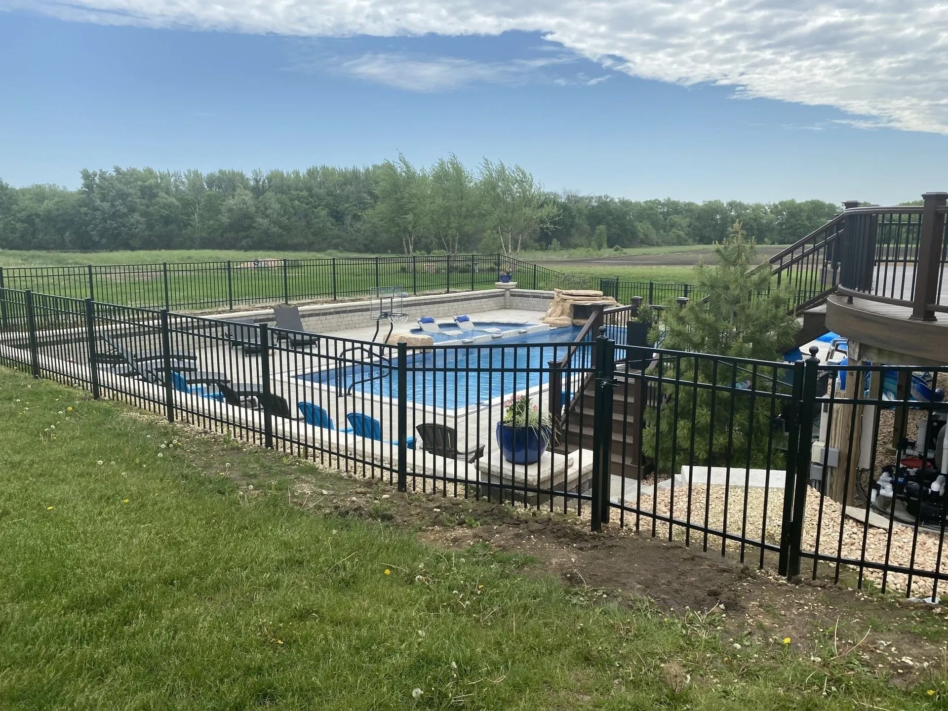 Black fenced-in outdoor pool on a green lawn, with a deck on the right and trees in the background under a blue sky.