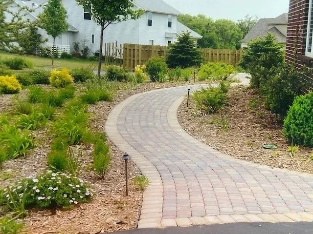 Brick pathway winding through a landscaped yard with various plants and houses in the background.
