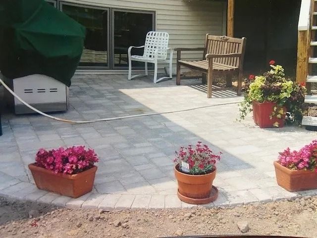 Patio with brick pavers, potted flowers, two chairs, grill, and a covered grill next to a house.