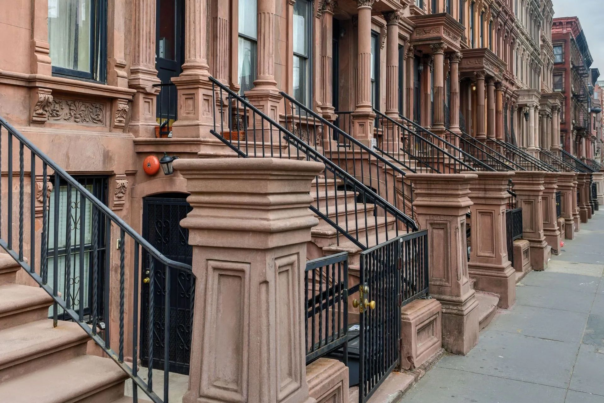 Row of brownstone stoops with black railings and stone columns along a city sidewalk