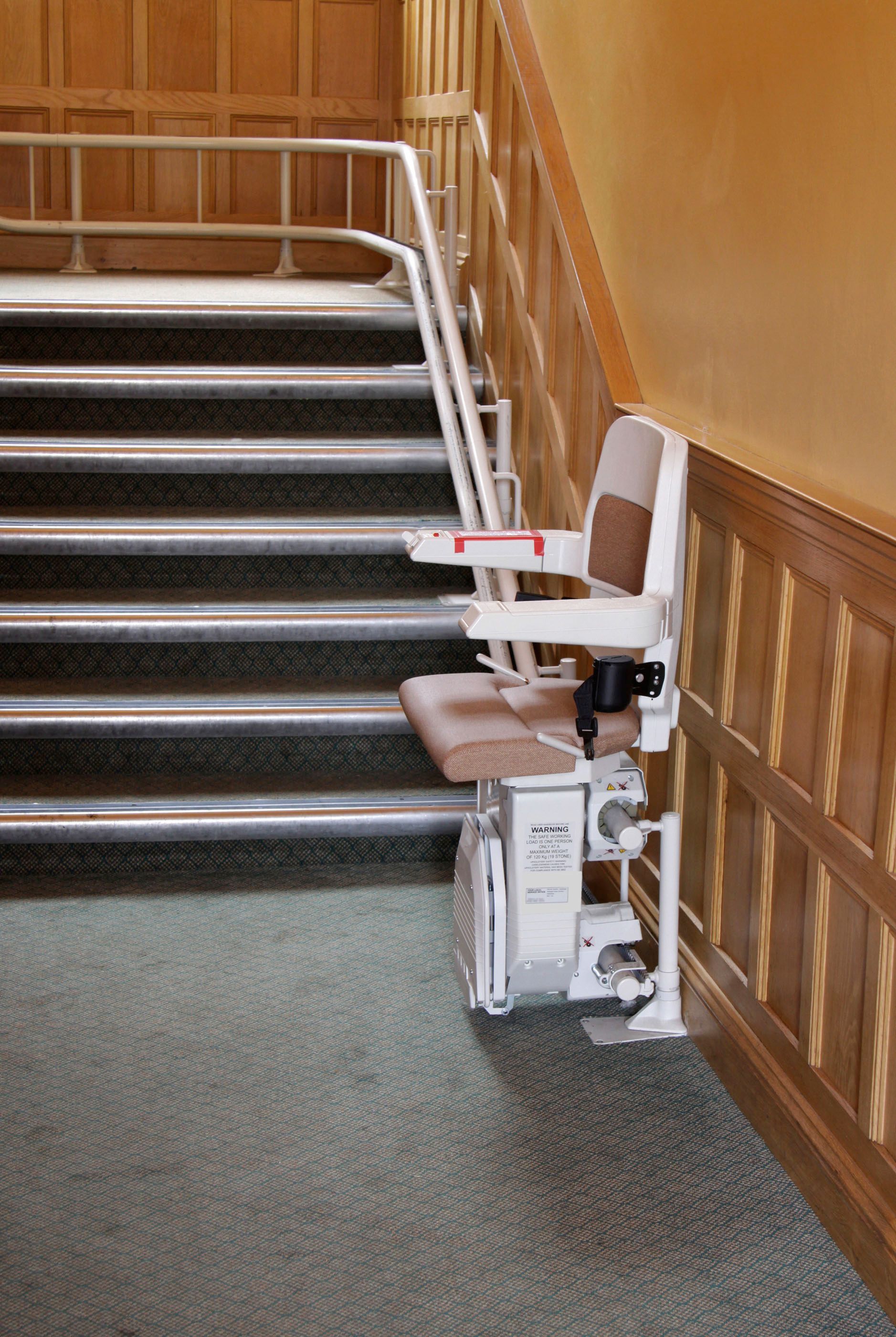 Stair lift on a staircase. Beige chair, white track, wood paneling, carpeted floor.