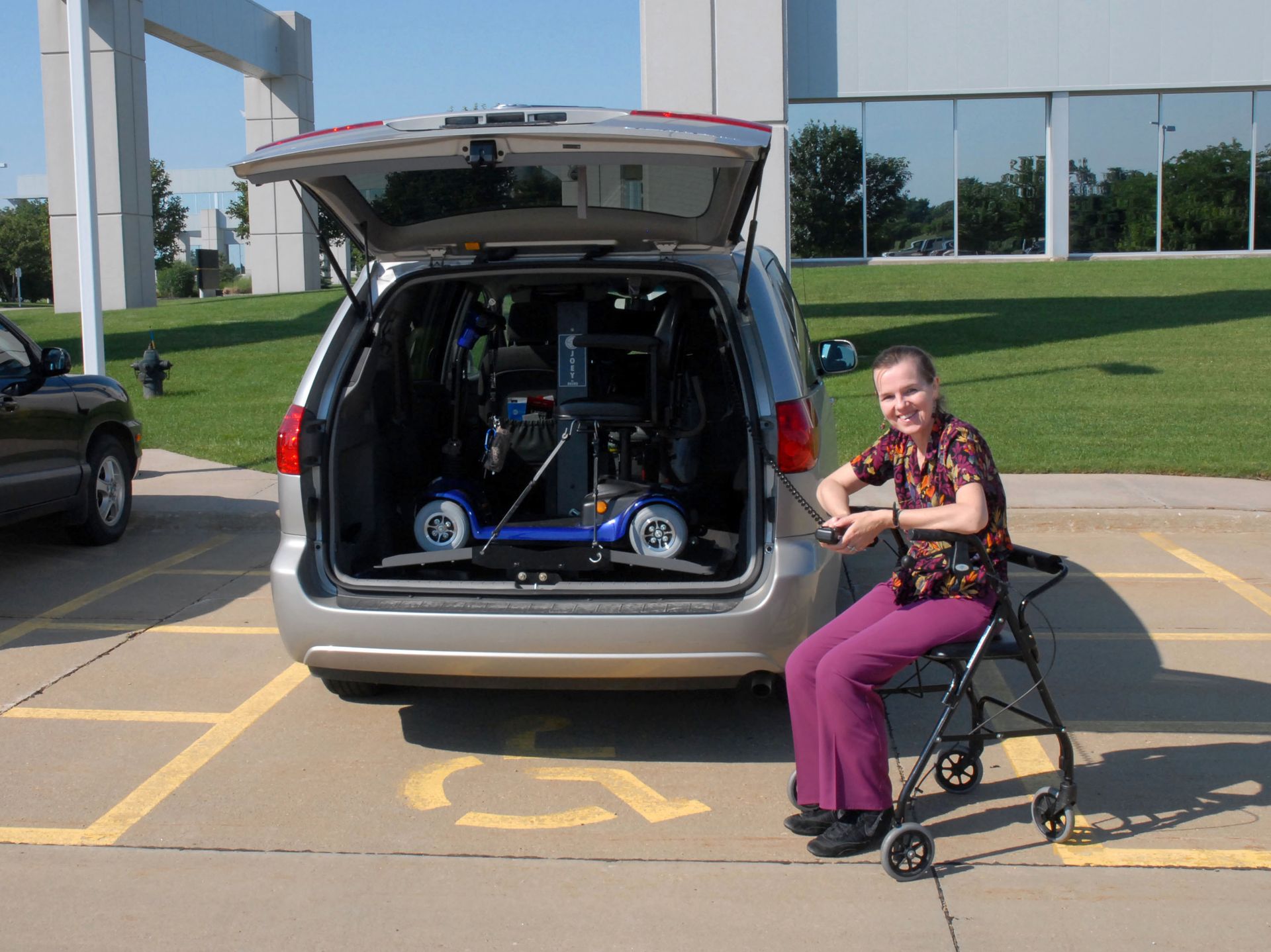Woman with a walker beside a van with an open back. A mobility scooter is inside. Parking space has accessible symbol.