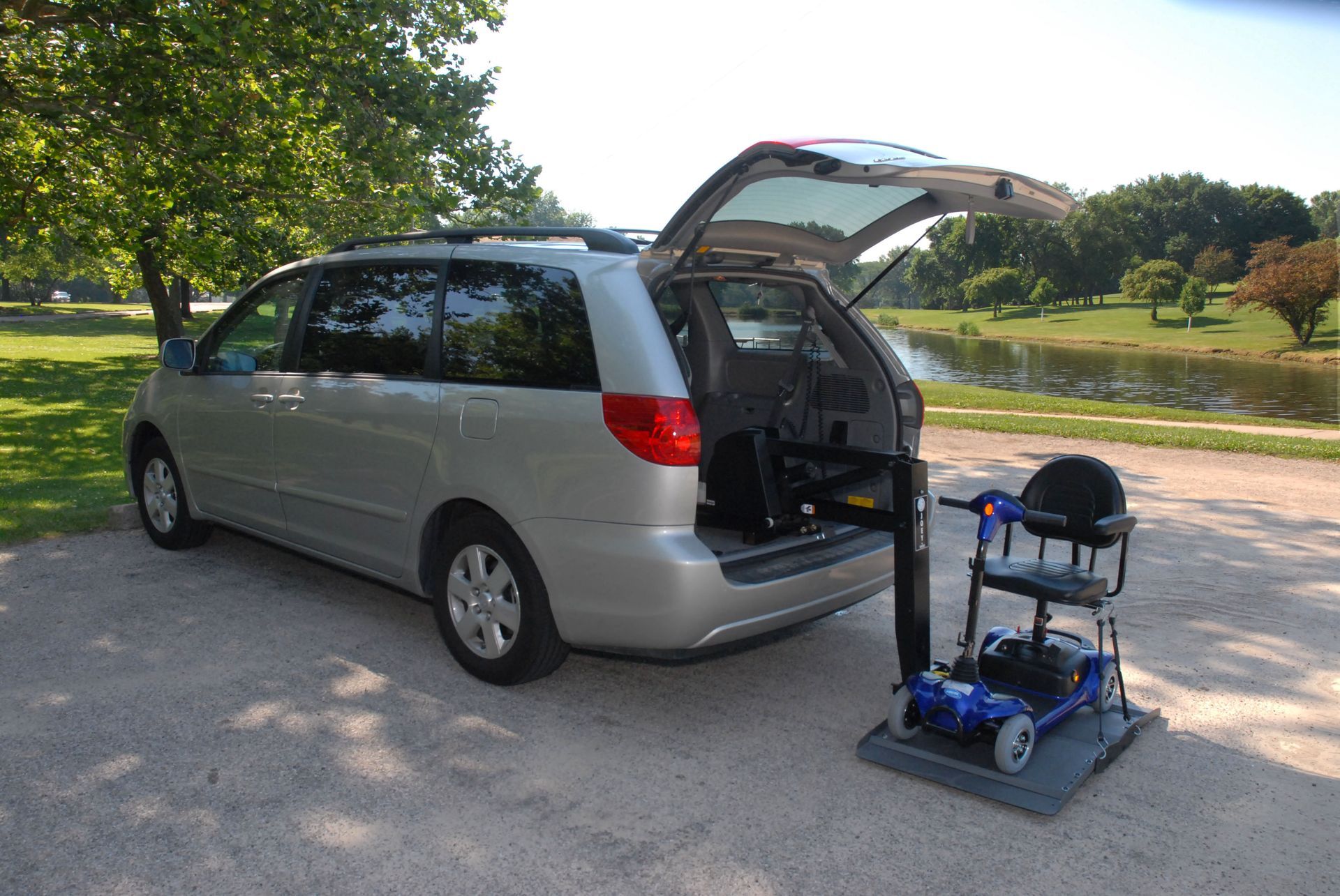 Silver minivan with lift and scooter on a gray platform. Outdoors by trees and a pond.