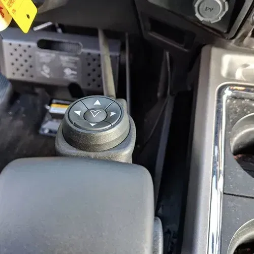 Black circular control knob with arrow buttons on a vehicle's console, near a cup holder.