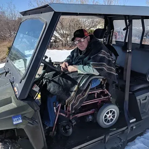 Person in wheelchair driving an all-terrain vehicle. Wrapped in blanket, sitting inside the cabin, outdoors on a sunny day.