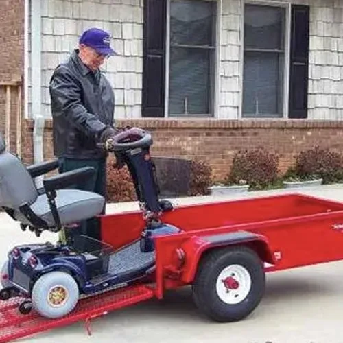 Man on mobility scooter loading it onto a red trailer ramp outdoors.