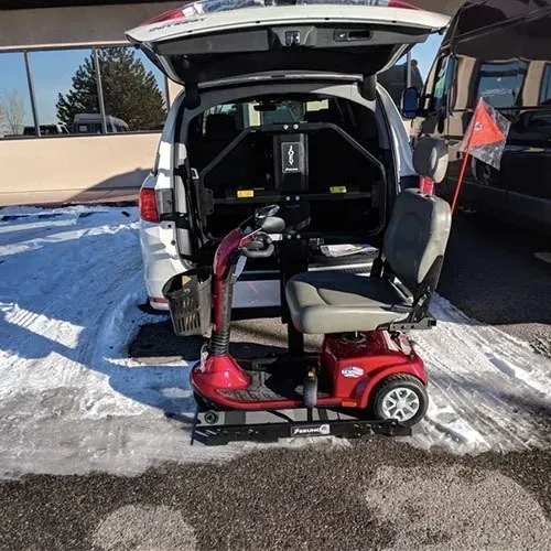 Red mobility scooter on a platform, loading into the open back of a white van, snowy ground.