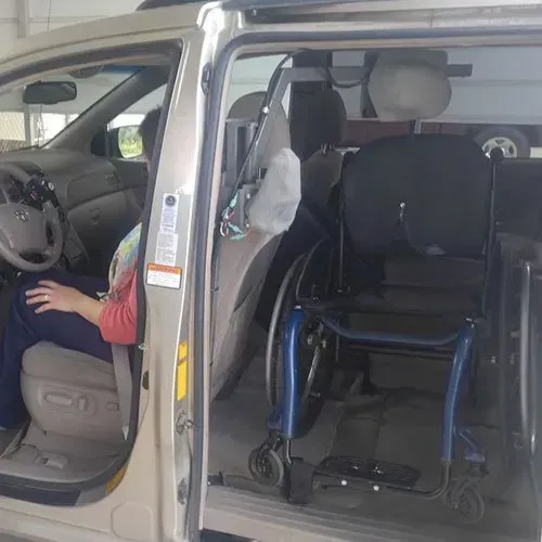 A person in a car, with the side door open. A wheelchair is next to the car seat.