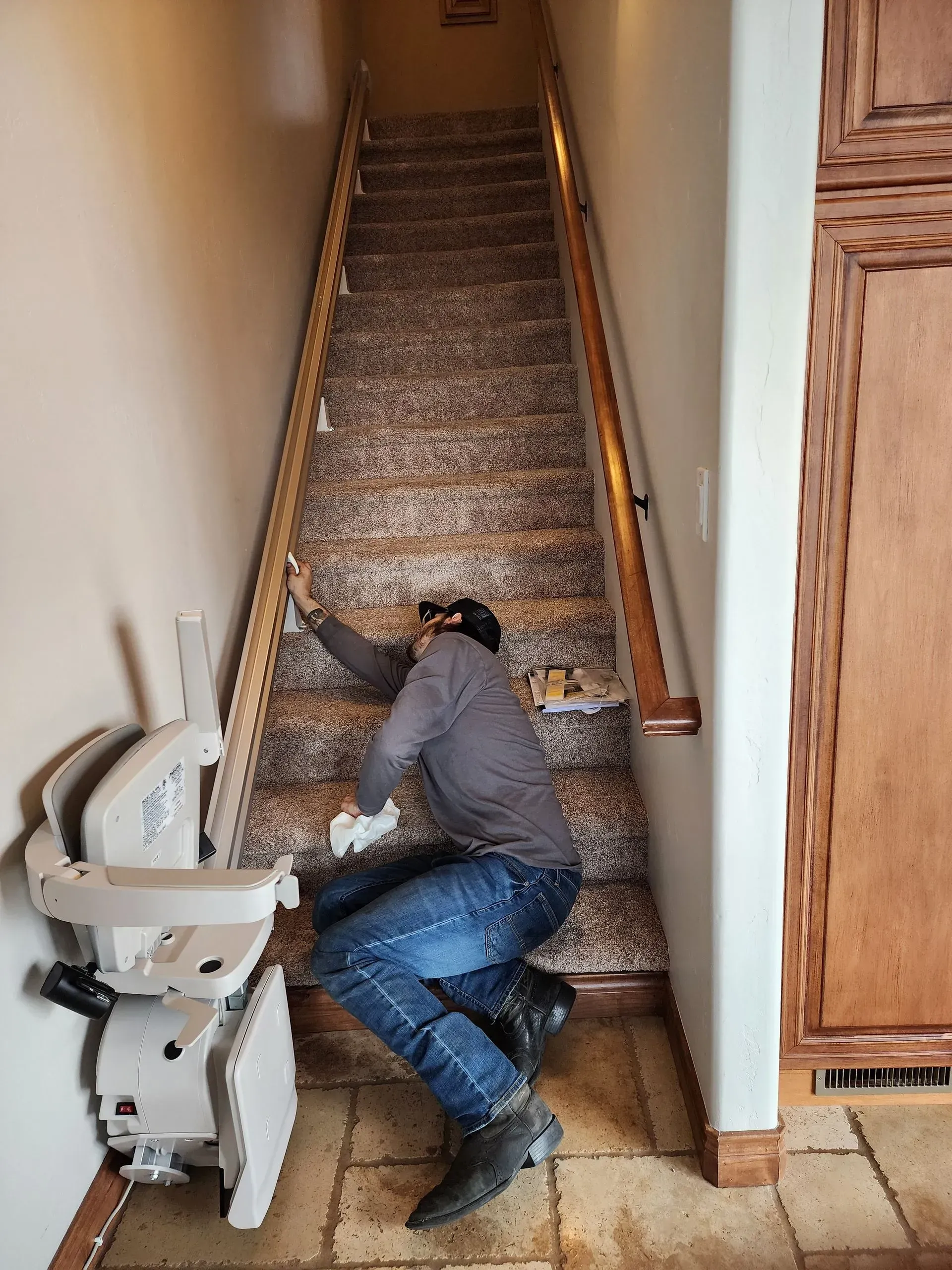 Man working on stair carpet next to stair lift; beige walls, brown door, tiled floor.