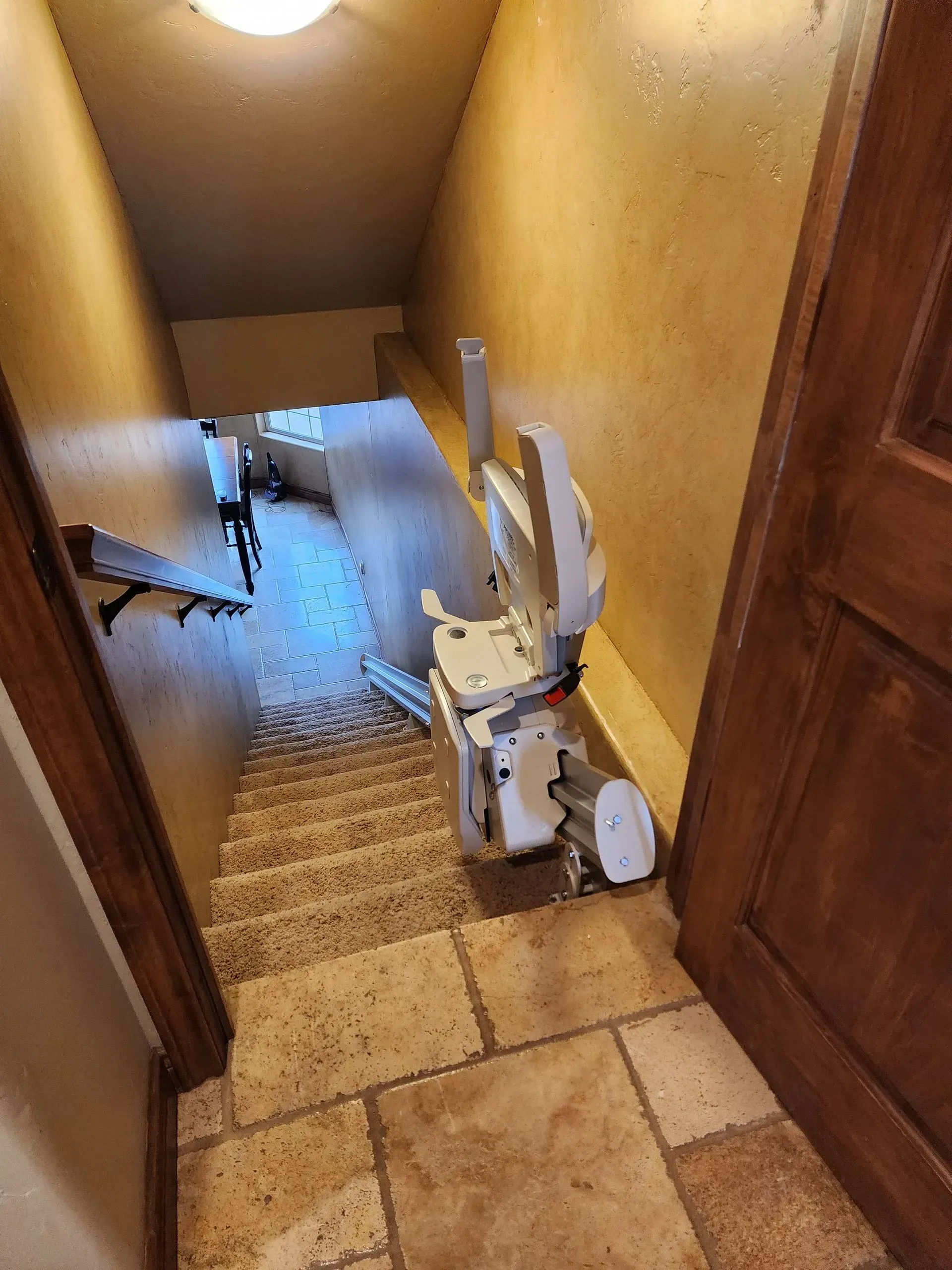 Stair lift installed on a narrow staircase with beige walls and stone tile flooring.