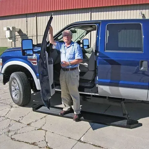 Man standing by open door of a blue pickup truck with a retractable step, near a building.