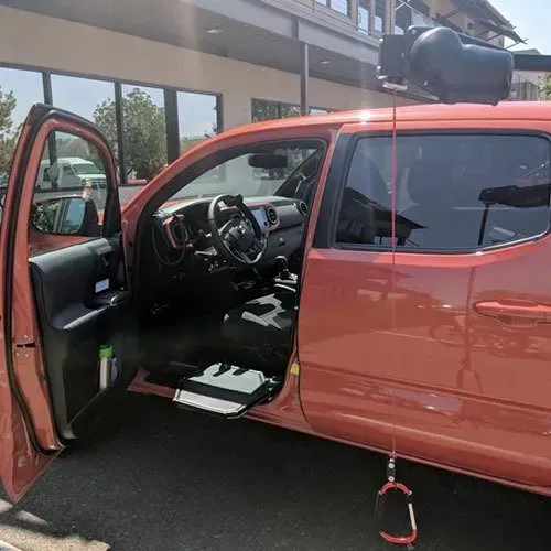 Coral-colored truck with open door; a camera rig is mounted on top; parked outside a building.