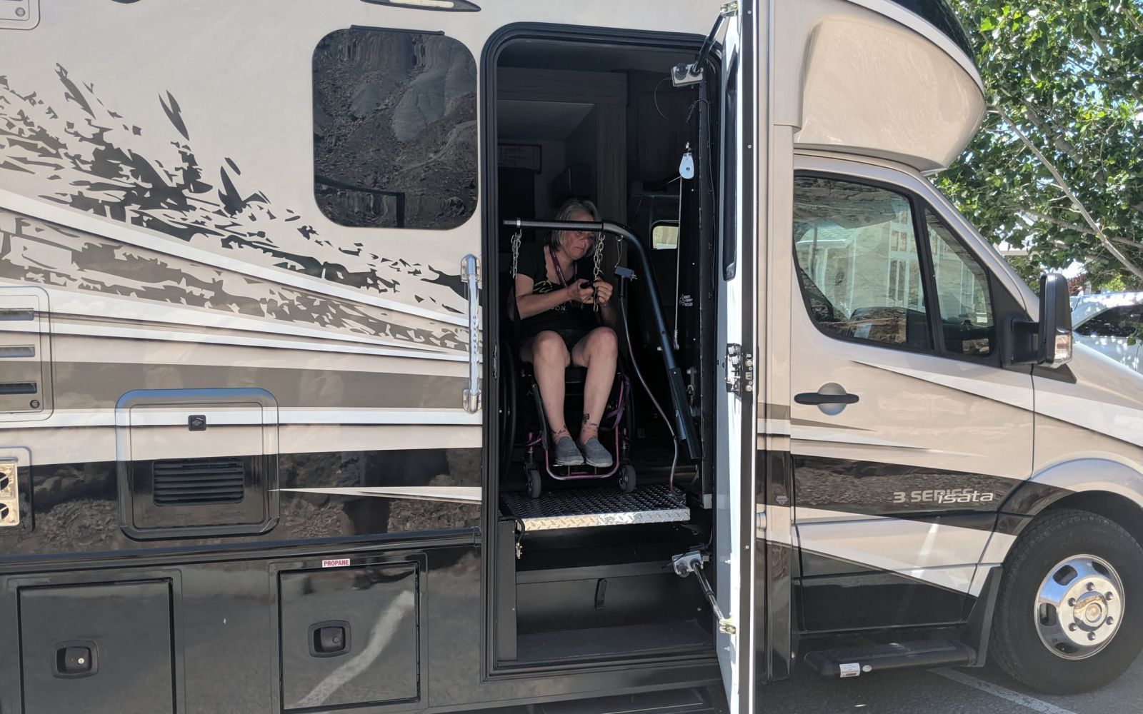 Person in wheelchair using lift to enter RV. Open door, parked outdoors, sunny.
