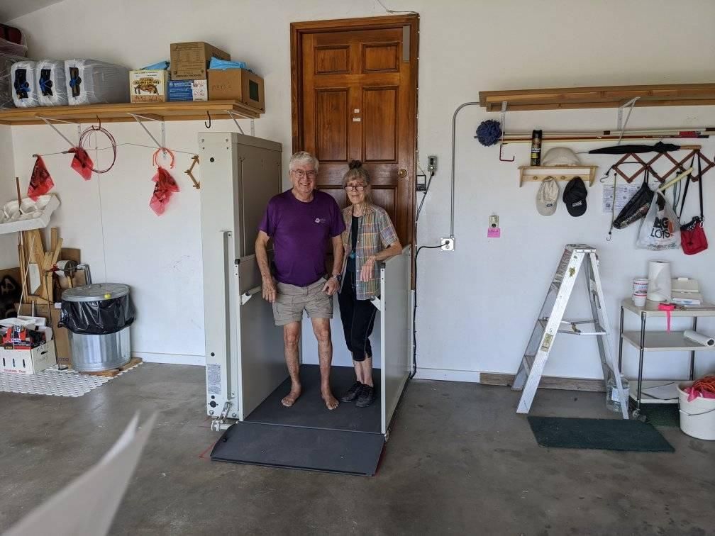 Couple standing in a garage lift. Man in purple shirt, woman in print shirt. Wooden door in background.