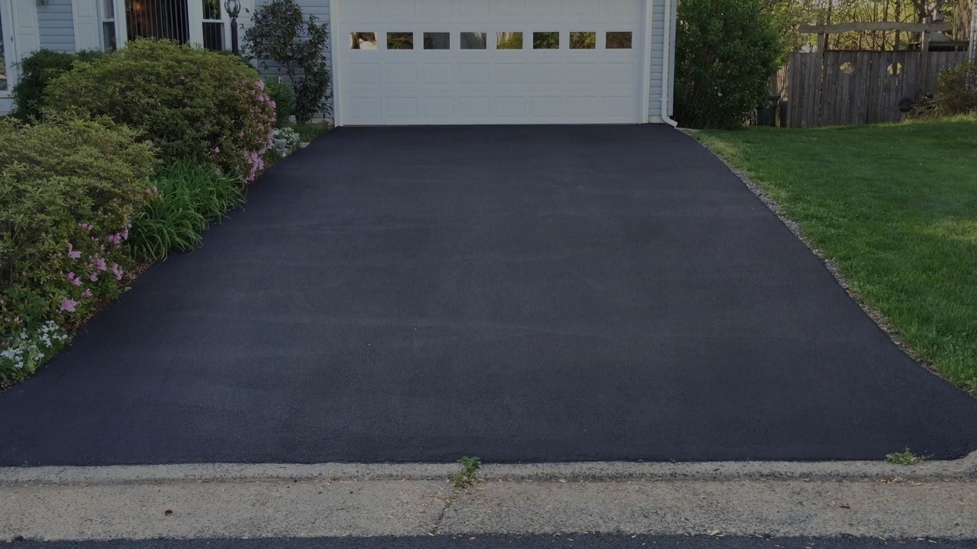 A freshly paved black asphalt driveway leads to a closed white garage door, flanked by bushes and a grassy lawn.