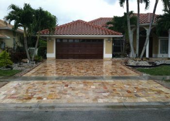 A residential house with a beige exterior, a terracotta tiled roof, and a tan stone-paver driveway leading to a garage.