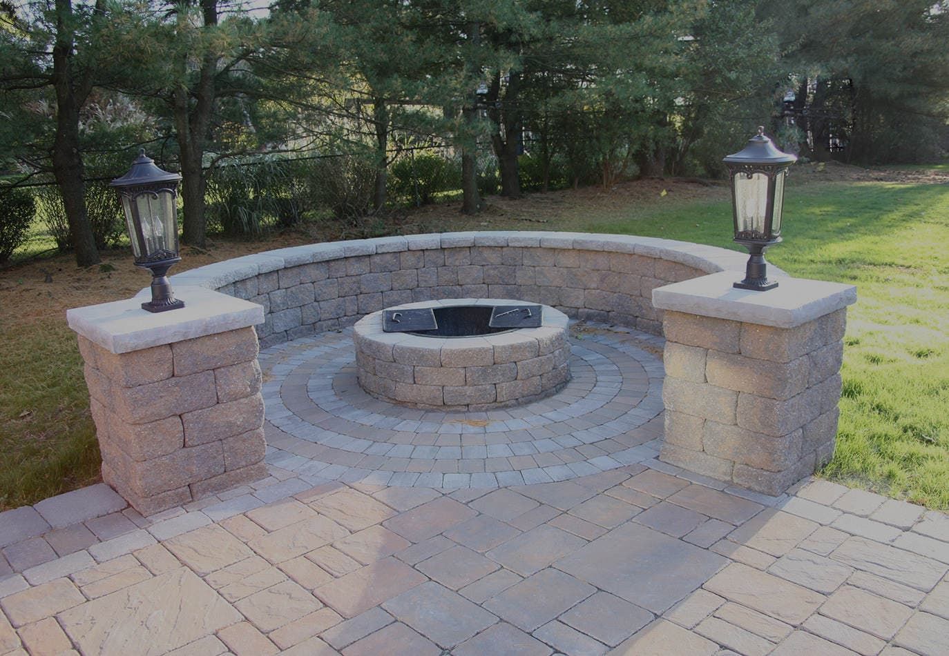 A semi-circular stone fire pit set into a brick patio, flanked by two stone pillars topped with lanterns.