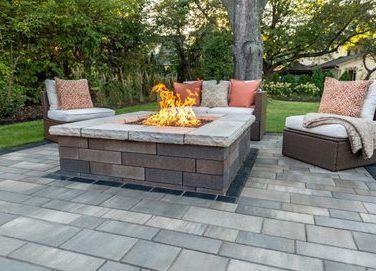 A square stone fire pit with a glowing fire on a paved patio with outdoor furniture set in a green backyard.