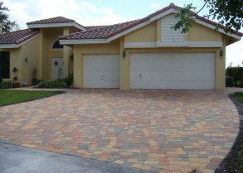 A yellow one-story house with a multi-car garage and a wide driveway paved with multicolored bricks.