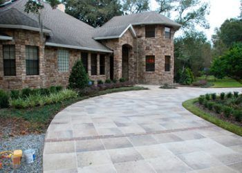 A multi-story stone house with a curved, light-colored stone paver driveway leading to the entrance.