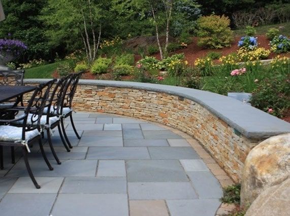 A stone patio with a curved retaining wall and seating area, featuring blue-grey pavers and a lush landscaped garden.