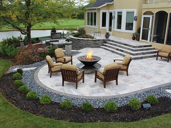 A stone patio featuring a central fire pit surrounded by five armchairs, set near a house and a landscaped backyard.
