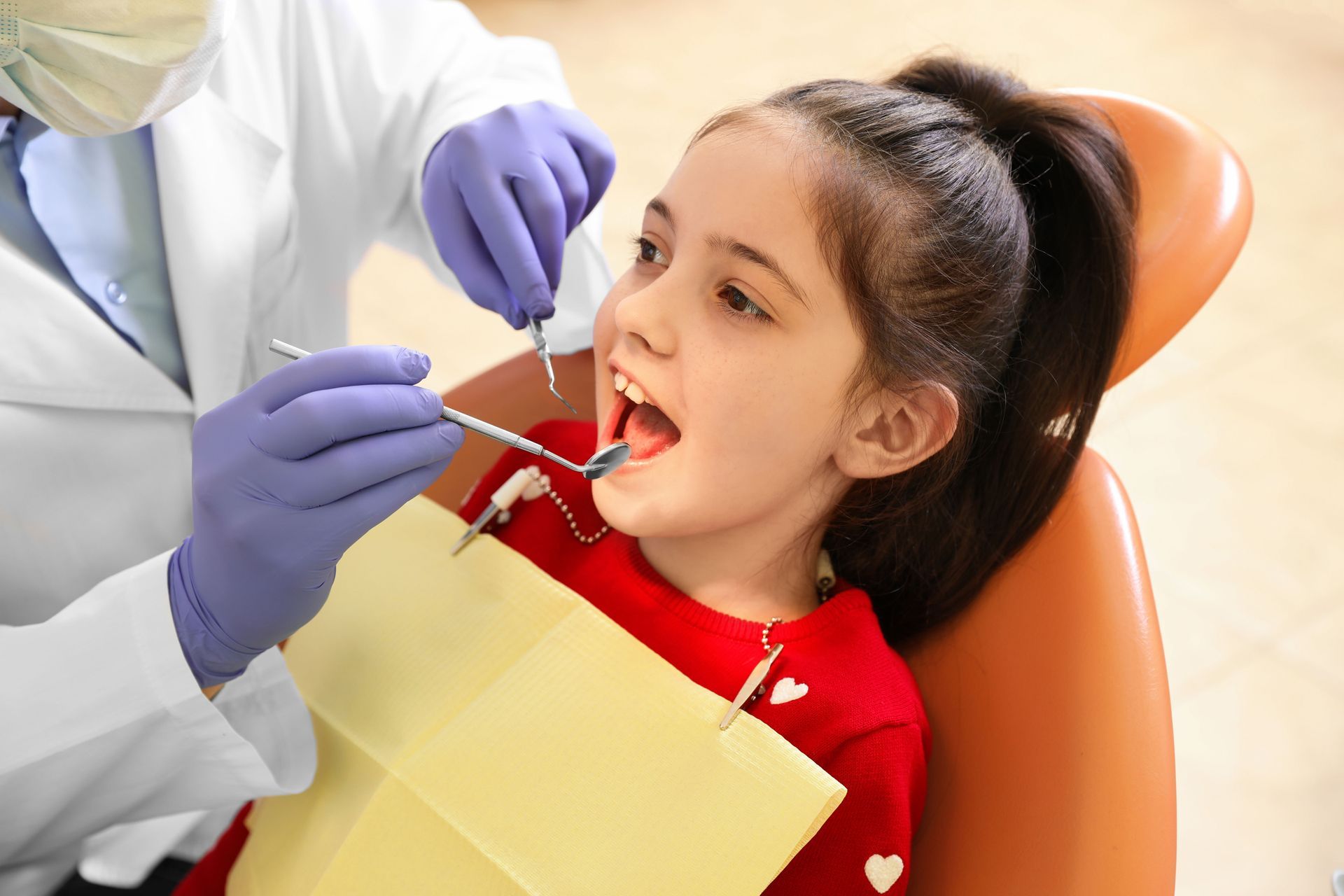 Dentist examining a child in an orange chair while holding dental tools in a clinic