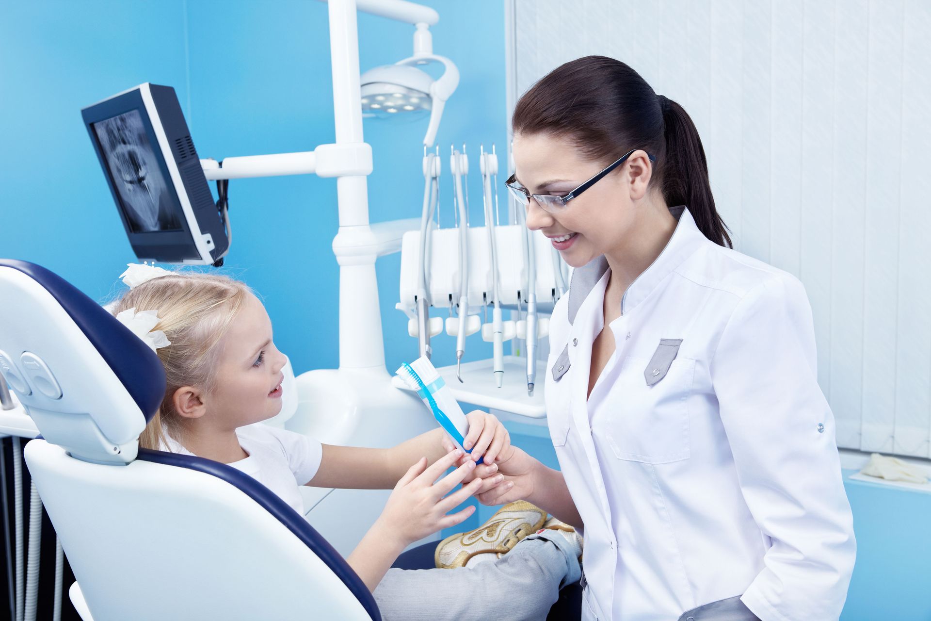 A dentist wearing a white coat hands a blue toothbrush to a patient sitting in a dental chair.