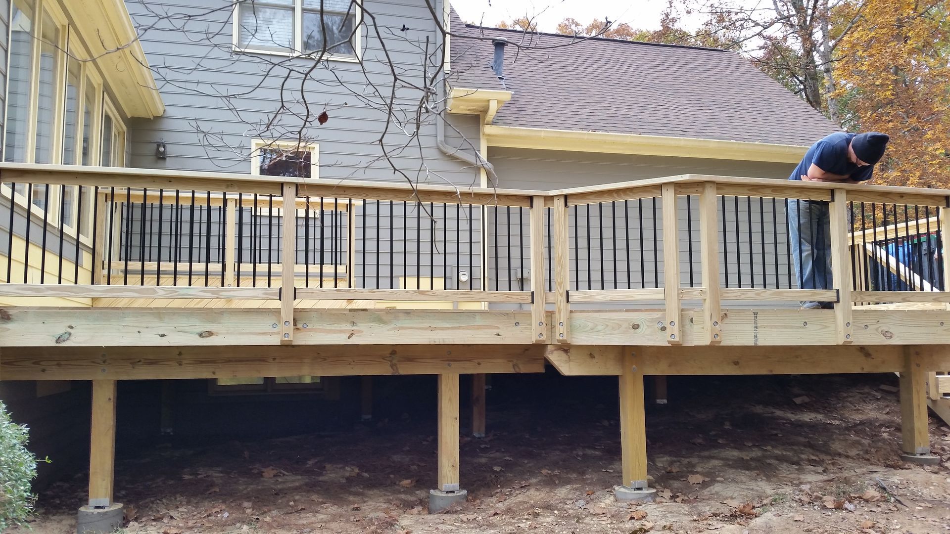 Man working on a wooden deck attached to a gray house; brown ground below.